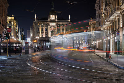 Illuminated street light trails on road by buildings in city at night