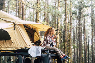 Full length of woman holding coffee cup sitting on motor home in forest