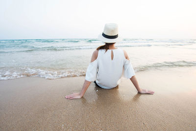 Rear view of woman sitting on shore at beach against sky