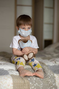 A little boy in a mask sitting on the bed and hug a teddy bear. image with selective focus