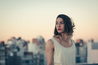 Thoughtful young woman looking away while standing against clear sky in city during sunset