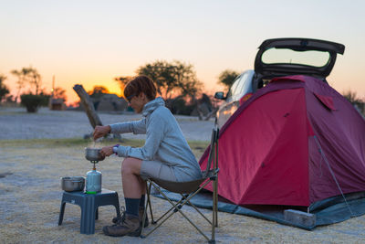 Woman cooking with camping stove while sitting by tent during sunset against sky