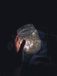 Cropped hand of woman holding jar with illuminated string lights on bed in darkroom
