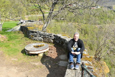 Portrait of man sitting on retaining wall against mountains