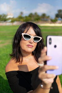 Woman wearing sunglasses making selfie on smartphone in sunlight over grass background.
