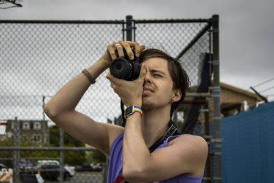 Young man holding camera while standing by fence against sky