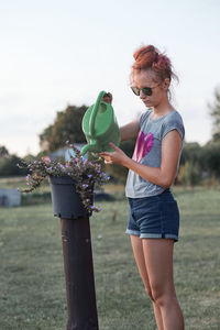 Girl watering plants while standing on grass against sky