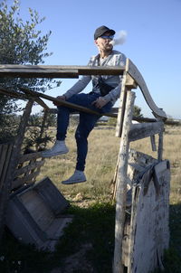 Man exhaling smoke while sitting on built structure against clear sky