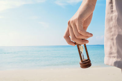 Cropped hand holding hourglass at beach against sky