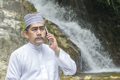 Thoughtful man talking on phone while sitting against waterfall