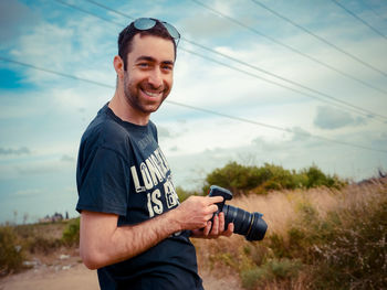 Smiling man holding camera while standing against sky