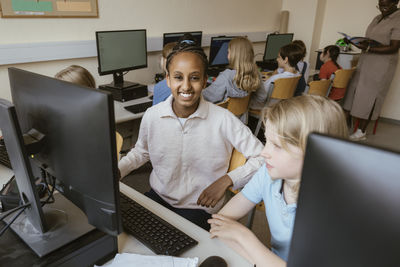 Portrait of smiling girl sitting on chair with friends in computer classroom at school