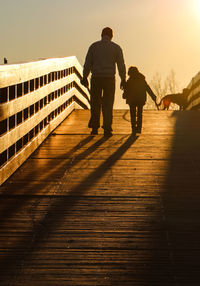 Rear view of man walking with son on bridge against sky during sunset