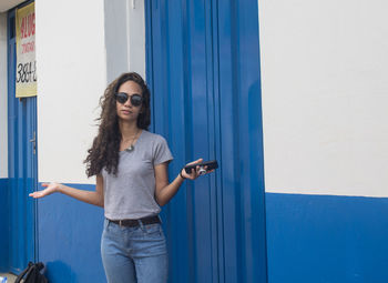 Young woman wearing sunglasses gesturing while standing against blue wall outdoors
