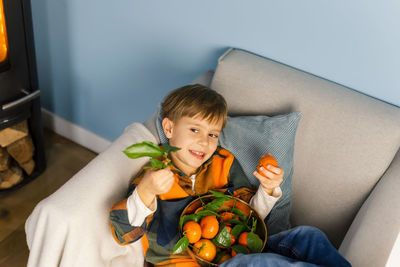 Boy peels and eats a tangerine, sitting in an armchair by the fireplace.