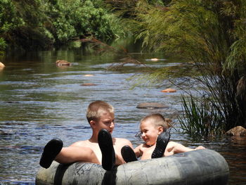 Shirtless brothers floating on inflatable ring over river against trees