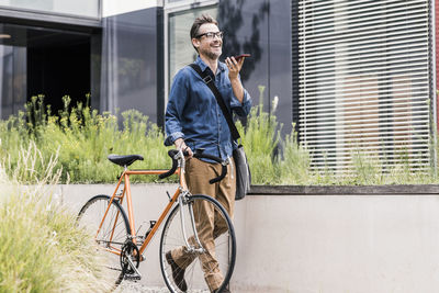 Smiling businessman with cell phone pushing bicycle