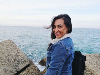 Portrait of smiling young woman standing at rocky beach against sky