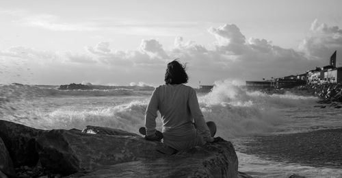 Rear view of woman sitting on rock by sea against sky