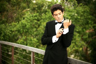 Handsome young man in suit buttoning cuff link while standing by railing against trees