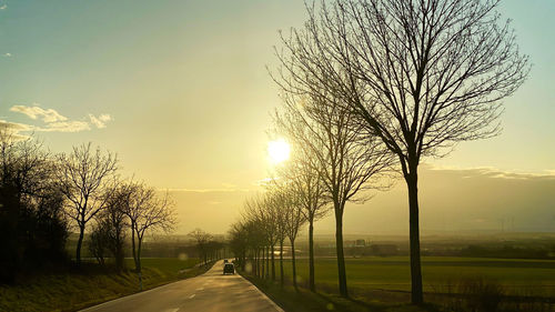 Road amidst bare trees on field against sky at sunset