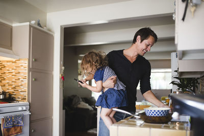 Daughter using phone while smiling father working in kitchen