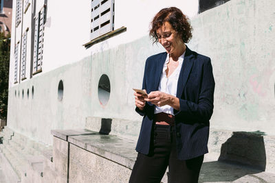 Positive adult female executive worker wearing classy outfit standing browsing on cellphone in sunny day