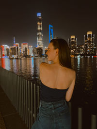 Young woman standing by railing against illuminated buildings in city at night