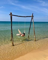 Rear view of shirtless mid adult man swinging at beach against blue sky