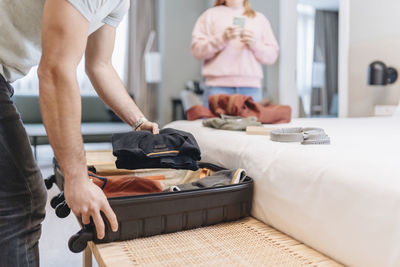 Man removing clothes from suitcase with woman in background at hotel