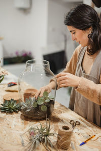 High angle of tranquil adult woman in casual clothes and gray apron planting small green succulents into glass terrarium while standing at table in contemporary light apartment