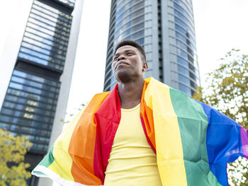 Happy young man wrapped in rainbow flag in front of skyscrapers