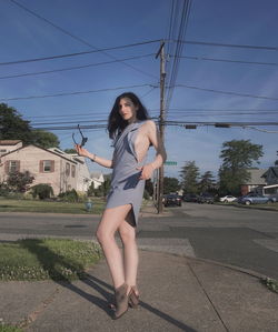 Portrait of young woman standing on sidewalk in city against blue sky