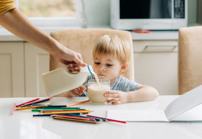 A little girl has breakfast in the kitchen and draws, her mother pours milk into a mug.