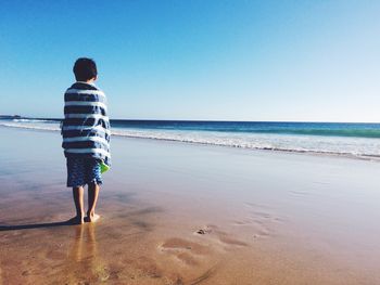 Rear view of boy wrapped in towel standing on shore at beach against clear sky