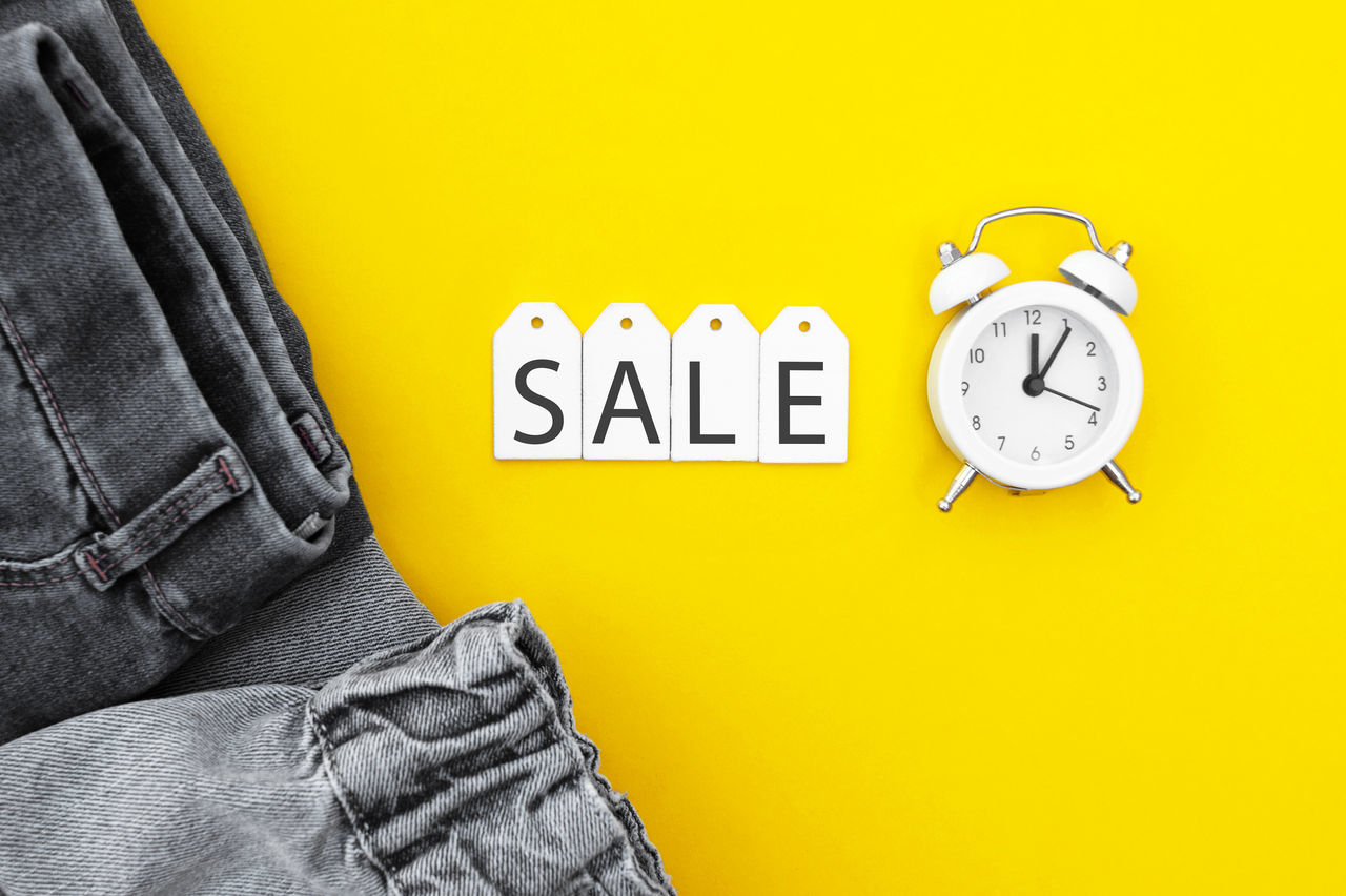clock, yellow, time, indoors, casual clothing, communication, clothing, text, number, jeans, one person, adult, font, wall - building feature, sign, men