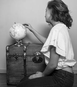 Side view of young woman holding globe on luggage at home