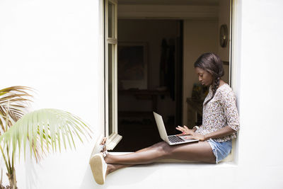 Full length of woman using laptop while sitting on window