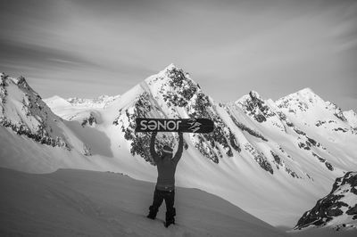 Rear view of man holding skateboard while standing on snowcapped mountain against sky