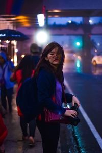 Portrait of young woman standing on footpath in illuminated city at night