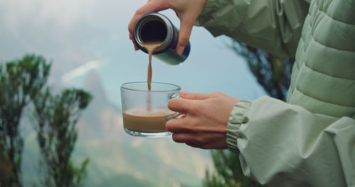 Female hands pour coffee from reusable container into a glass cup on nature mountain background.