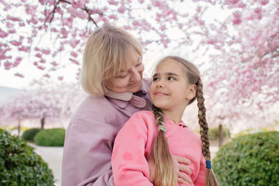 Girl hugs her grandmother near blooming branch of sakura during walking in spring garden