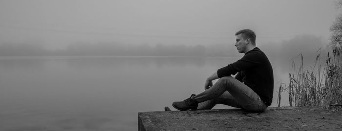 Young man sitting by lake against sky