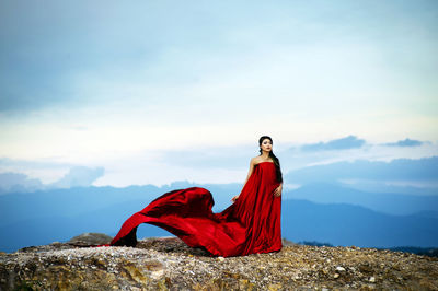 Female model in red dress standing on cliff against sky