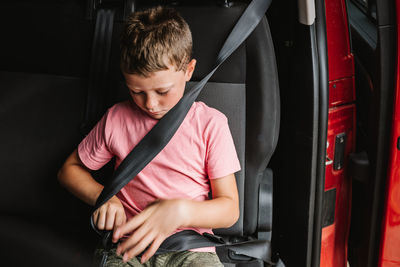 Unemotional boy in casual clothes buckling up while sitting on seat inside of car on back seat of car during road trip