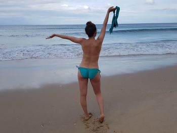 Rear view of shirtless woman holding bra while standing at beach