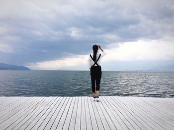 Full length rear view of woman in suspenders standing on pier against sea and cloudy sky