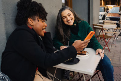 Happy african american female with curly hair sitting at table with dessert and showing cellphone to mature hispanic female in outdoor cafe