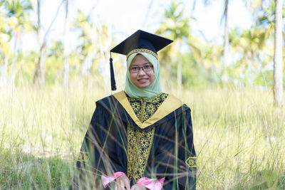 Portrait of young woman wearing hijab and graduation gown sitting amidst plants on field at park