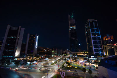 Illuminated city street by buildings against sky at night